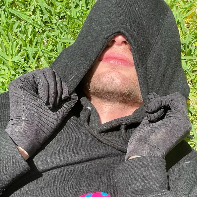 The first photo of Manatreed. He is lying in the grass, wearing a black hoodie and black gloves. He has pulled the hoodie up over his face to cover everything except the nostrils of the nose, the lips, and a scruffy chin. The face underneath is light-skinned and the hair is too patchy to definitevely say what color it is.