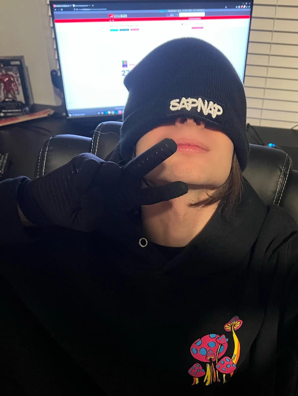 The third Manatreed photo. He's sitting in his office, turned away from his computer to the camera holding up a peace sign. He's still wearing a black hoodie and black gloves but is now also wearing a black Sapnap beanie that covers most of his face, leaving only the nostrils, mouth, and clean-shaven chin. His long brown hair pokes out from under the beanie, and on the computer screen in the back, we can see his youtube social blade stats. This is clearly a different person than the first set of photos.