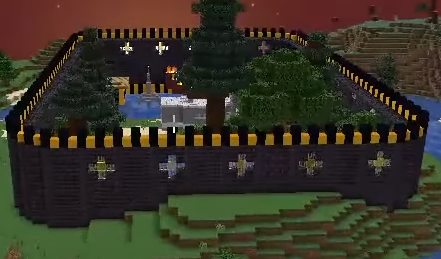 This is a screenshot of Minecraft gameplay. It shows the L'manberg walls surrounding the van, a pond, a few pine trees, and a couple of wooden buildings. The walls themselves are very tall, crafted from black bricks, and topped off with a yellow and black concrete rampart. Near the top of the walls are windows in the walls in the shape of a plus, but there are bars preventing people from entering that way. The rest of the background is Minecraft terrain with no additional builds.
