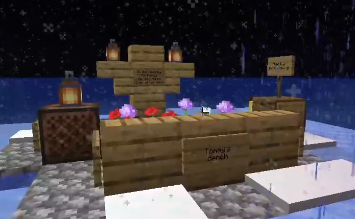 A screenshot of Tubbo's stream. It's his memorial that he made Tommy in Snowchester. There's a replica of Tommy's bench, a jukebox, a chest, and a sort of wooden statue to act as a grave marker. Beneath the statue, there are alliums and roses planted. There are signs on the objects, labeling them Tommy's bench, and explaining what the statue is, although in the picture it's too pixelated to make out. It's snowing.